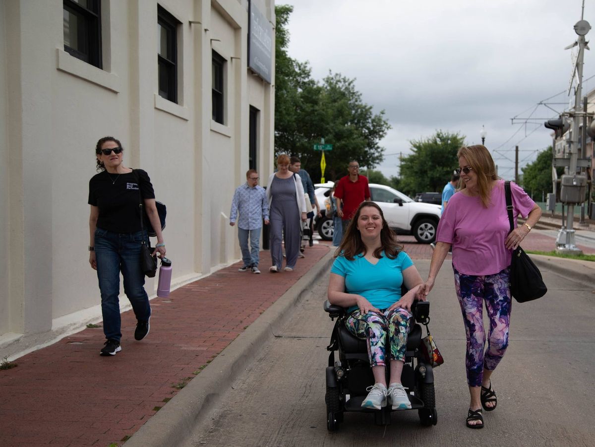 Friends walk down a city sidewalk, one in a power wheelchair.